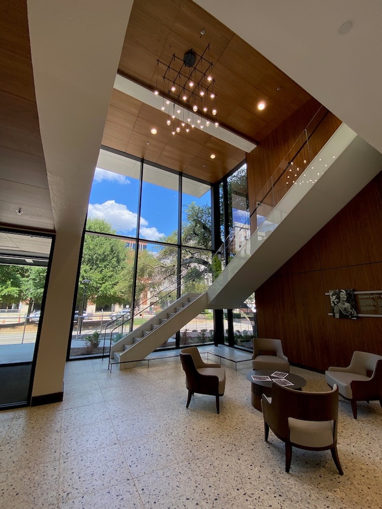 Inside of lobby facing floor to ceiling glass walls looking at University Boulevard. Shows terrazo floors, a seating area with four low chairs, a gorgeous pendant chandelier, and you see the open stairwell that leads to the second floor.