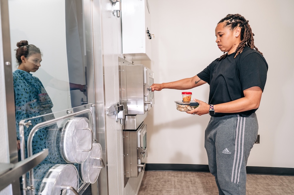On the left is a patient inside a calorimeter room. On the right a researcher has food in one hand and is reaching for the small door to deliver food to the patient.
