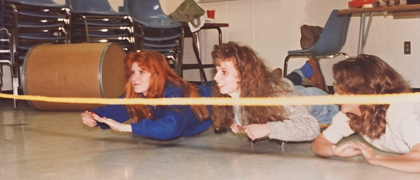 Three young women from the 1980s lying on their stomachs on the floor in front of a ribbon stretched before them.