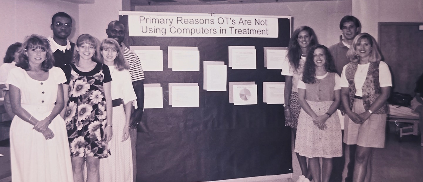 1990s image of students standing in front of a display reading "Primary Reasons OTs are not Using Computers in Treatment." Picture is black and white.