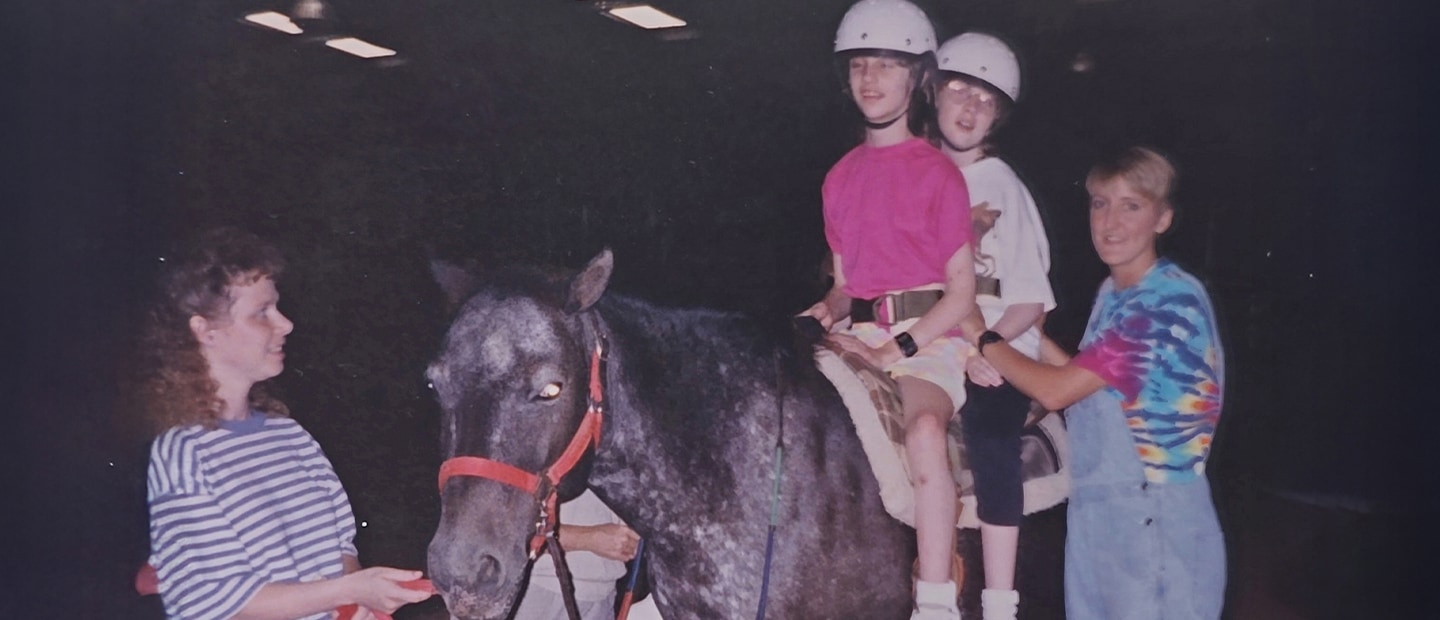 Two children in safety helmets ride a horse,  accompanied by two adult women.