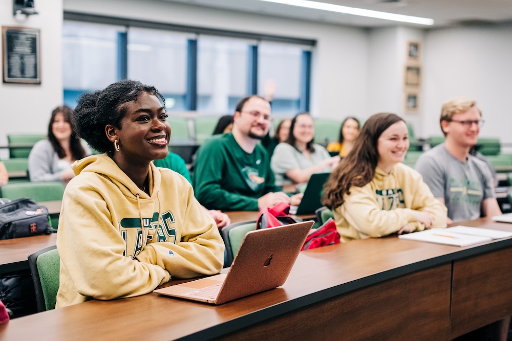 Smiling students are sitting at tables in a classroom listening to a class lecture.