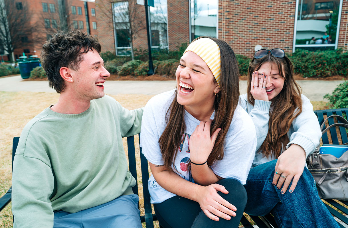 Three students laughing while sitting on a bench