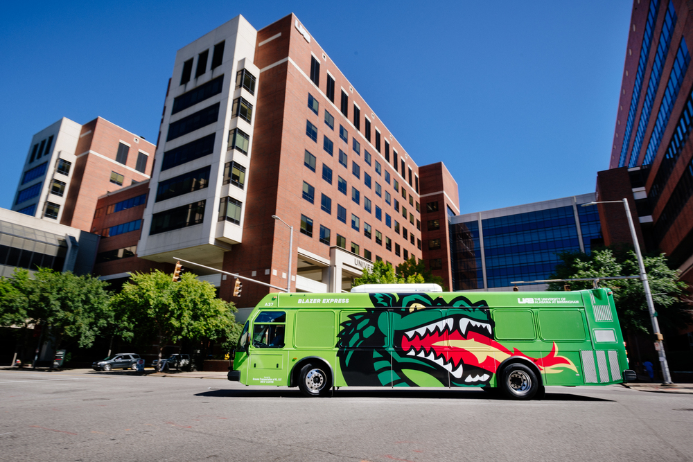 From side, a Blazer Express bus showing a graphic of Blaze (mascot) breathing fire traveling on 19th Street South in front of the North Pavilion of UAB Hospital.