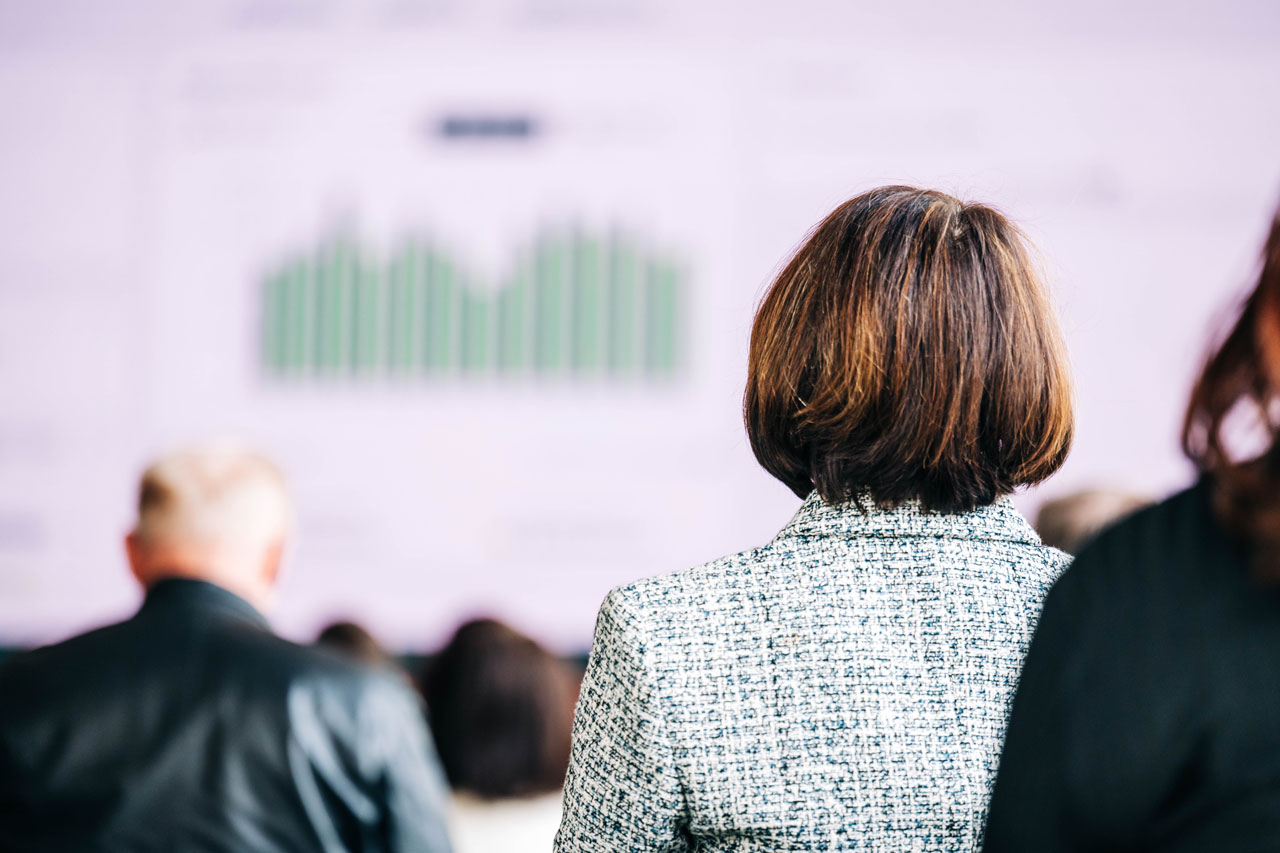 Audience members listen to a presentation with a chart projected on a screen.