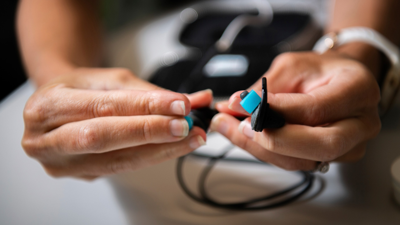 Closeup of hands holding black taVNS earbuds with blue hydrogel inserts on the portion that is inserted into the user’s ear canal.